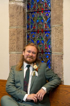 A groom in a sage green suit poses next to a stained glass window in a church setting.