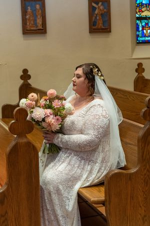 A bride in a white lace gown and veil sits in a wooden church pew holding a pink floral bouquet.
