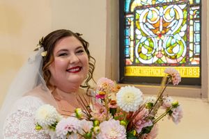 A bride in a white lace dress holds a bouquet of pink and white flowers in front of a stained glass window.