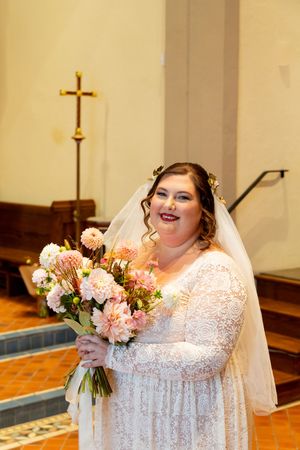 A bride in a lace wedding dress holds a pink and peach bouquet while posing in a church.