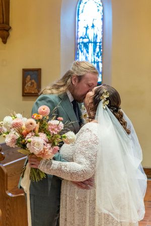 A couple embraces while holding a pink and peach floral bouquet in a church.