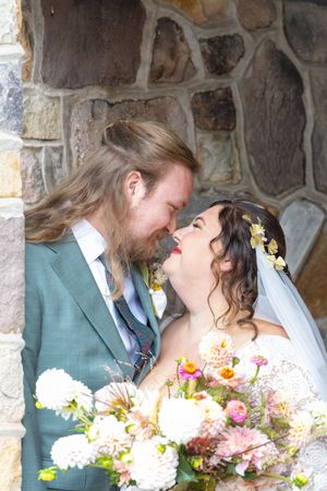 A joyful wedding couple shares a laugh while holding a colorful bouquet against a stone wall backdrop.