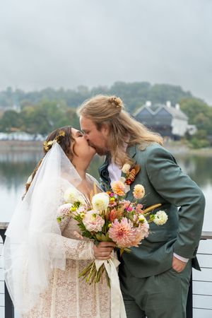 Newlyweds share a romantic kiss by a waterfront with autumn foliage in the background.