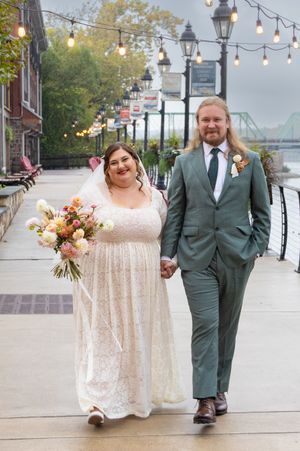 Wedding couple walks along riverfront boardwalk with string lights overhead and bride holding colorful bouquet.
