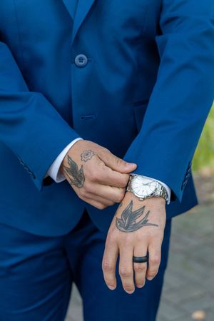 Close-up view of hands wearing a blue suit jacket showing tattoos and silver watch details.