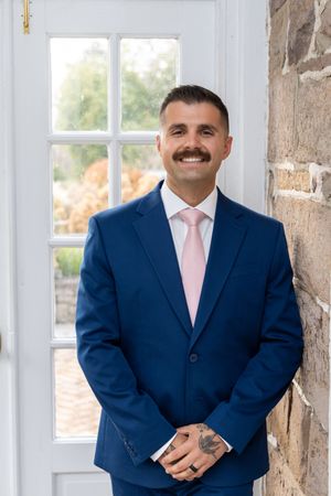 A person in a navy blue suit and pink tie smiles while standing against a stone wall with white-framed windows.