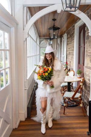 A bride in a white dress and cowboy boots twirls playfully in a rustic sunlit hallway with wooden floors and windows.