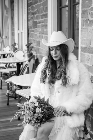 A bride in a white dress and hat holds a lush floral bouquet against a rustic brick wall backdrop.