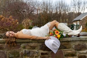 A bride in a white dress and cowboy boots lies on a stone wall holding a colorful autumn bouquet.