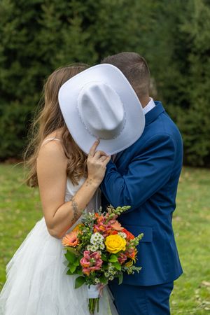 A couple shares a romantic moment behind a white cowboy hat while holding a vibrant bouquet at an outdoor wedding.