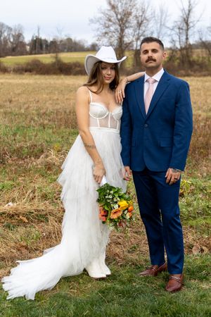 A couple in wedding attire poses together in a rustic field during autumn with sunflowers and greenery.