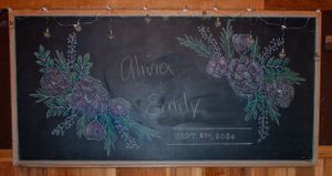 Wedding chalkboard sign decorated with floral illustrations in purple and green chalk.