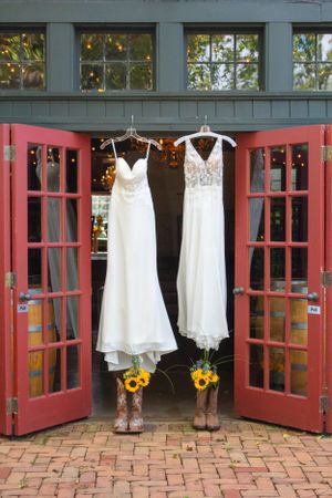Two white wedding dresses hanging in doorway with cowboy boots and sunflowers below.