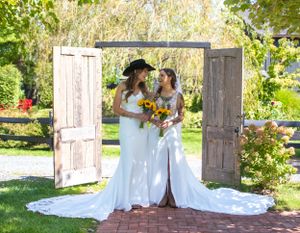 Two brides in white dresses stand together in front of vintage wooden doors in a garden setting.