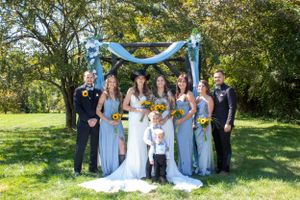 Wedding party poses under blue arch with sunflower bouquets in outdoor garden setting.