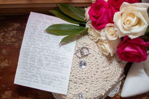 Wedding rings on lace doily with handwritten note and pink and white roses.
