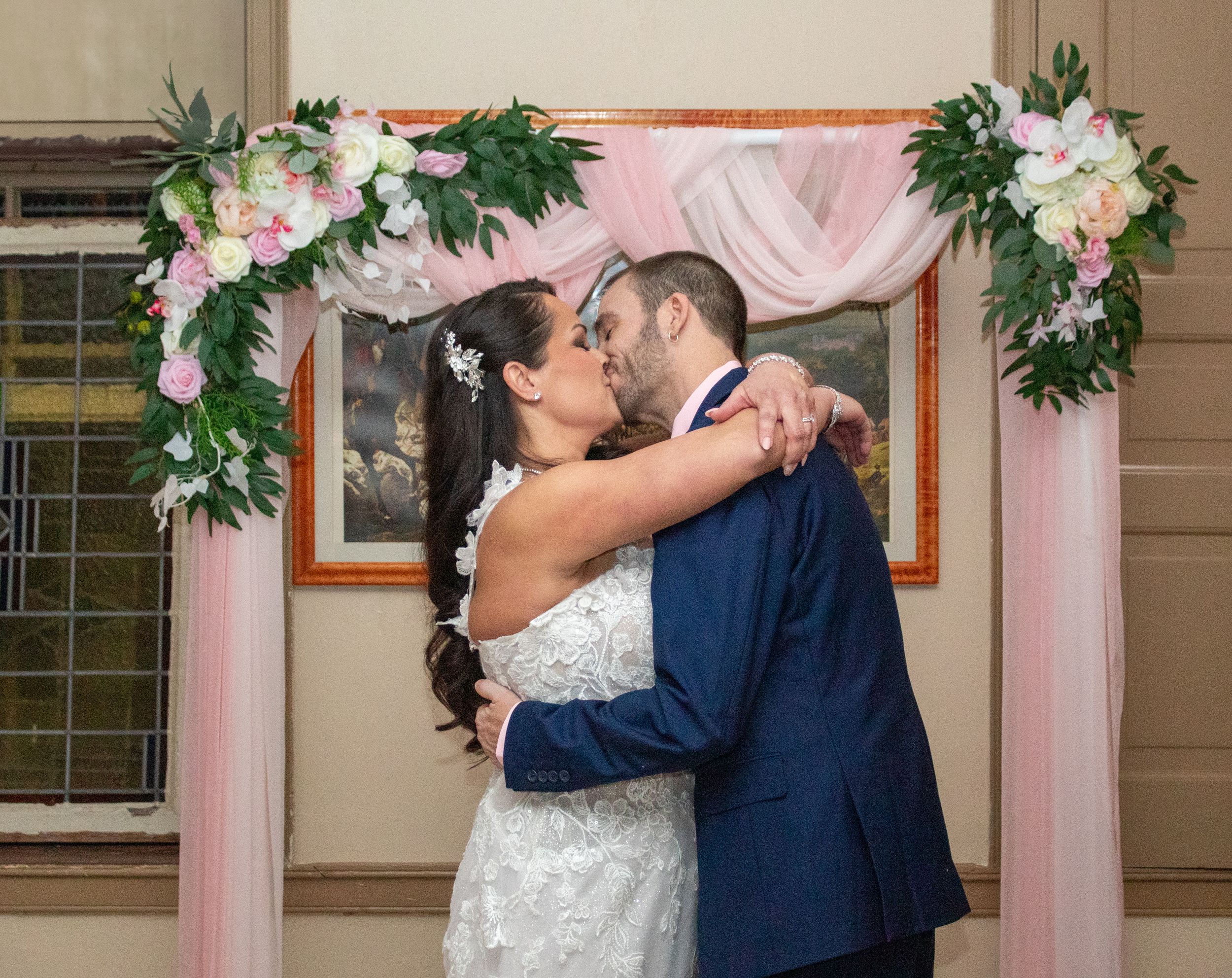 A wedding couple shares a kiss under a floral arch decorated with pink fabric and white flowers.