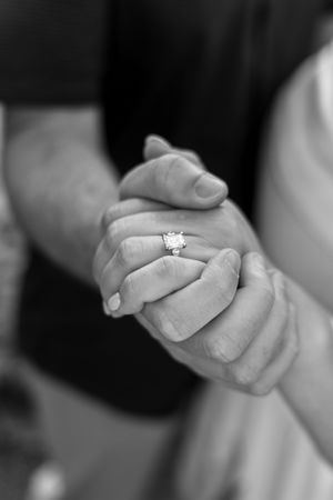Black and white photo of couple holding hands showing diamond engagement ring, romantic wedding moment.