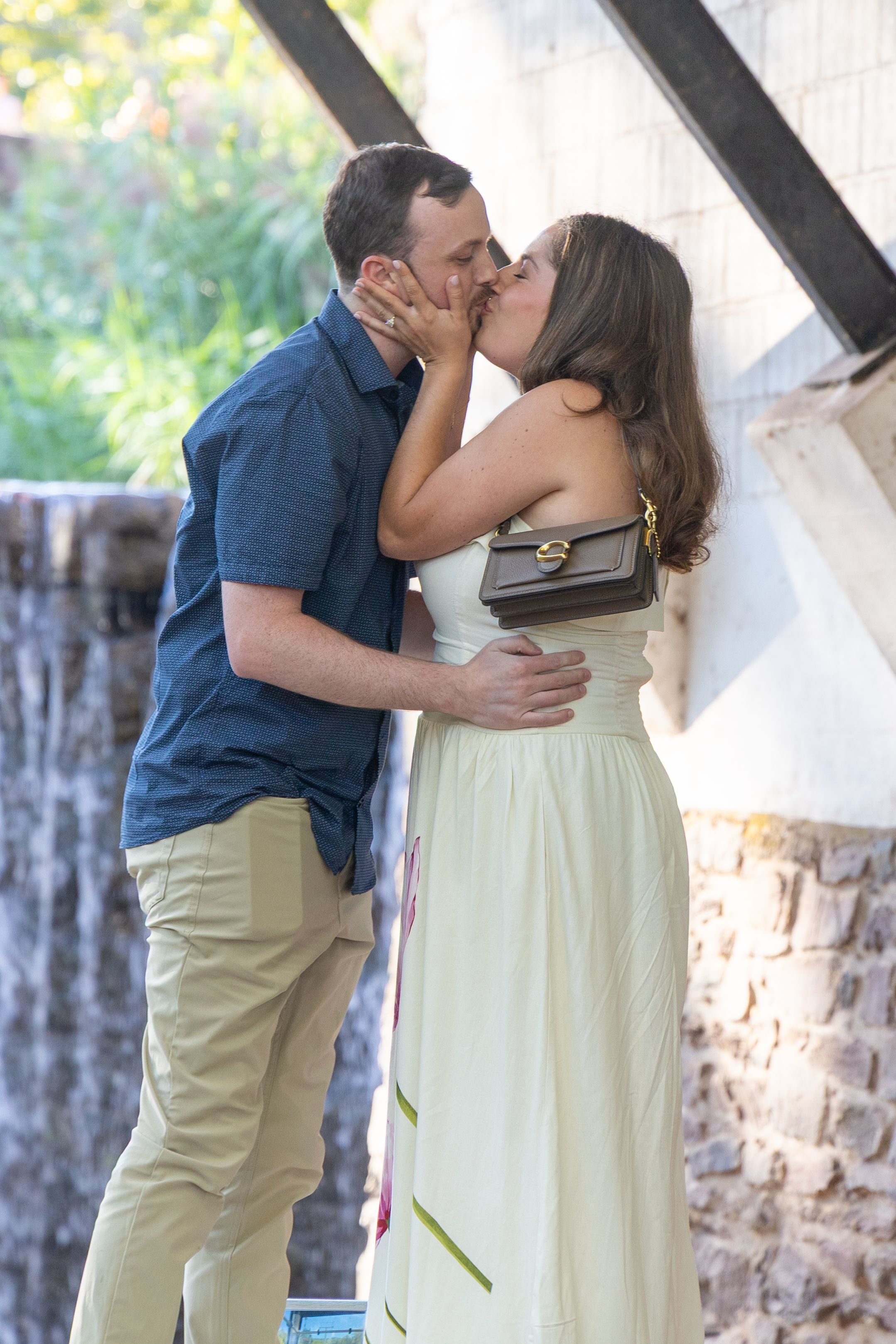 A couple shares a romantic kiss outdoors, woman in white dress with small purse, man in blue shirt and khaki pants.
