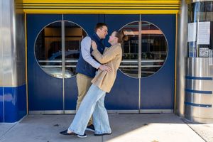 Two people dance together in front of a building entrance with oval windows and blue walls.