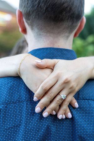 Close-up of a woman's hand with an engagement ring and gold bracelet resting on a man's shoulder in a blue shirt.