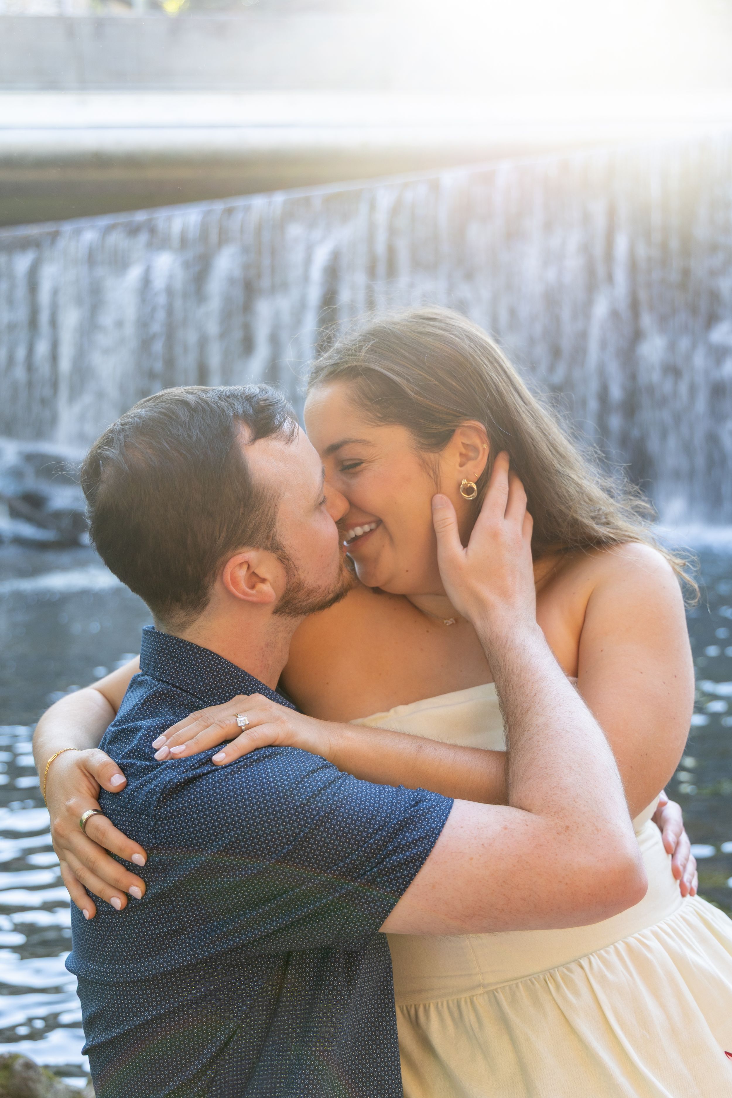 Couple sharing a romantic kiss in front of a sunlit waterfall, woman in cream dress with engagement ring visible.