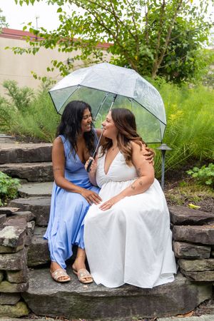 Two women in blue and white dresses share a clear umbrella while sitting on stone steps in a lush garden.