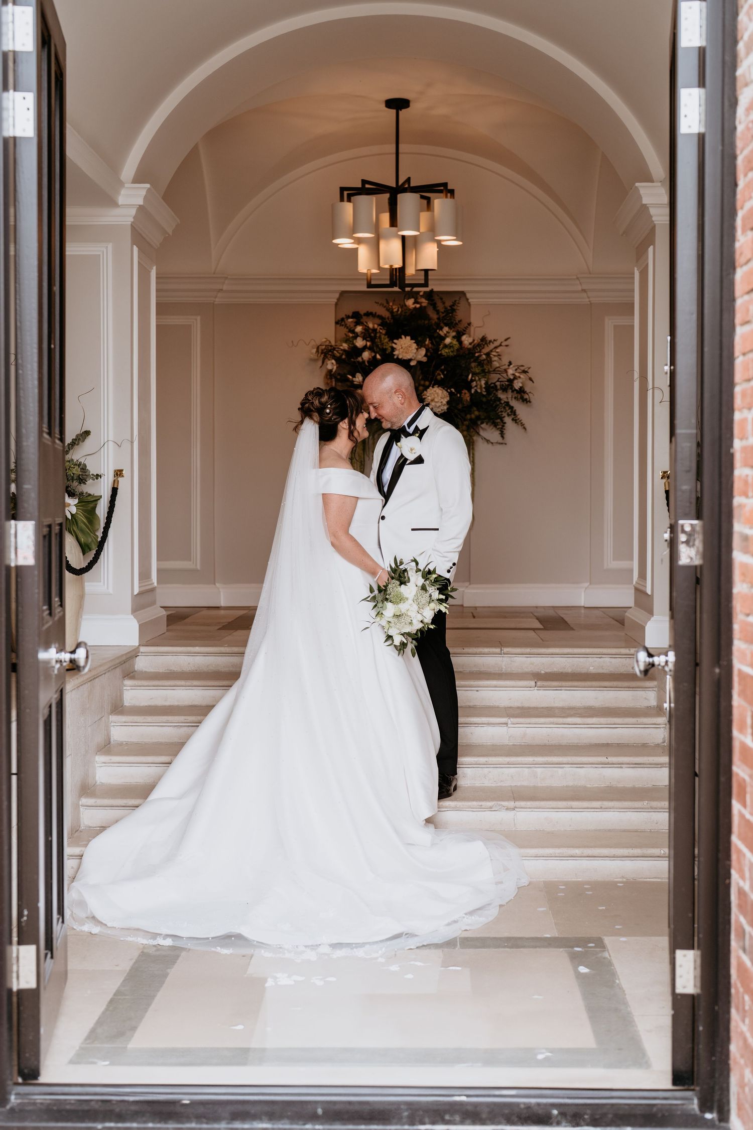 Wedding couple sharing an intimate moment in an elegant arched hallway with chandeliers and white decor AT SANT FFRAED HOUSE