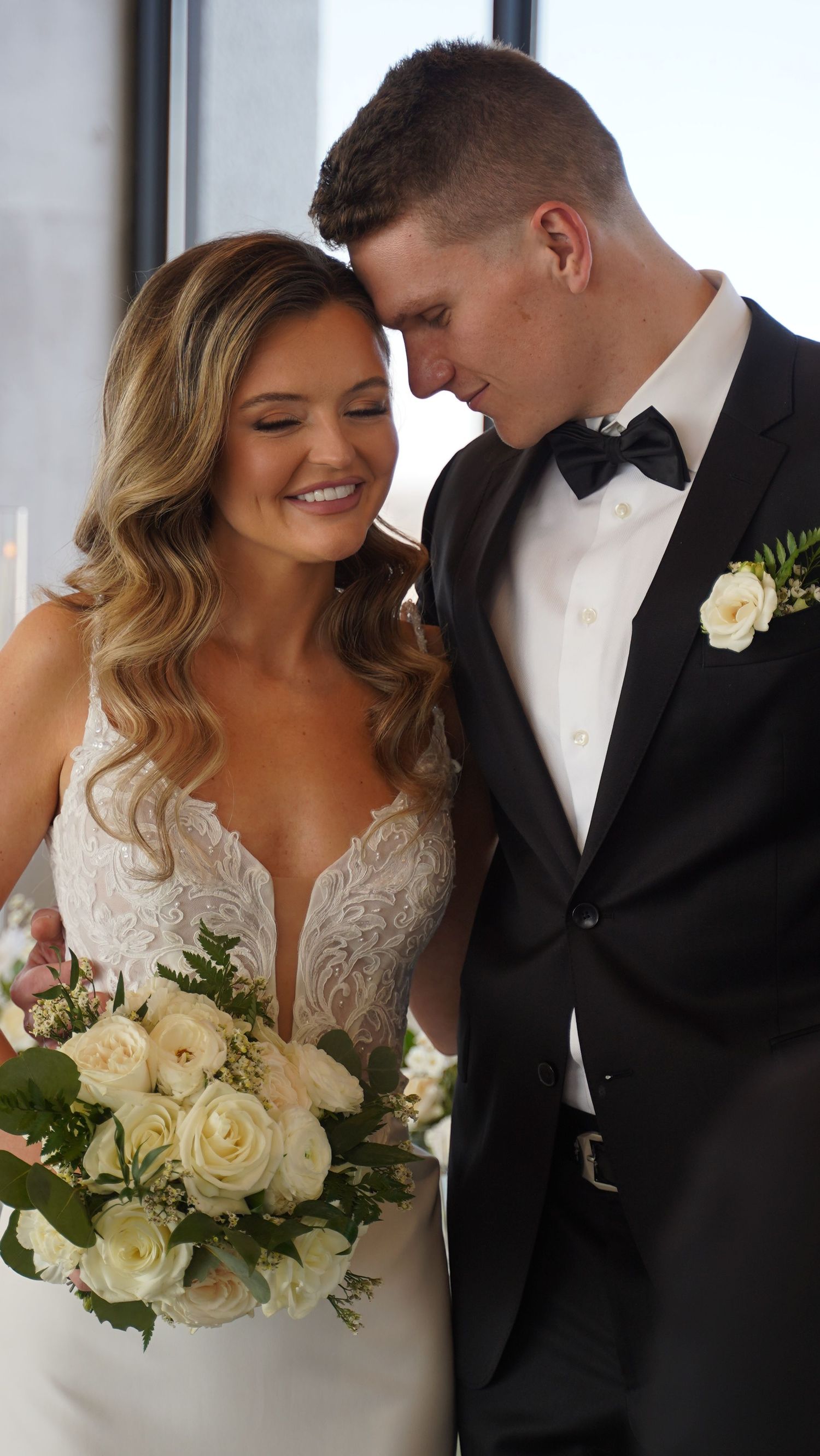 A tender wedding moment captured with a bride in a lace dress holding white roses and groom in classic tuxedo.