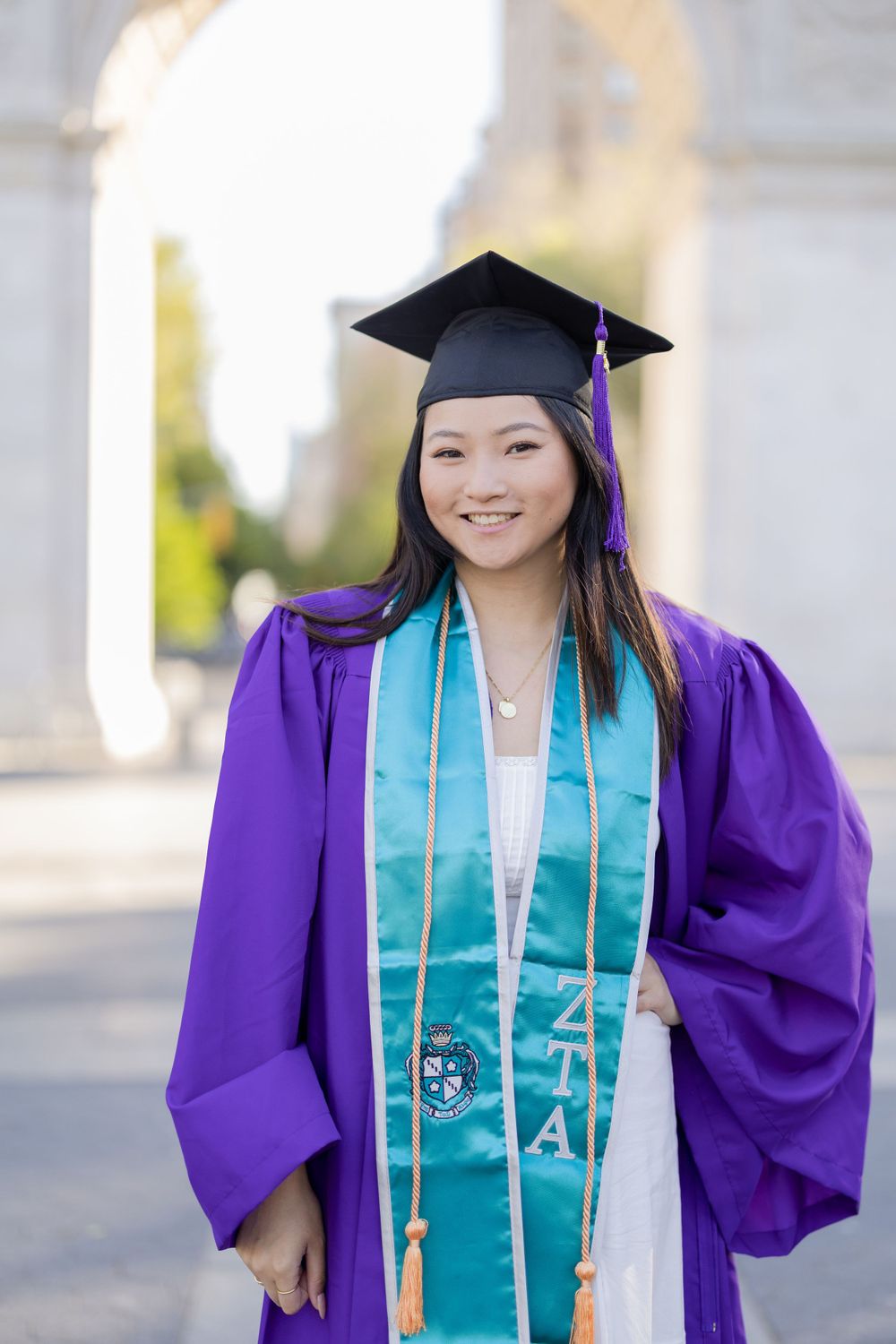 Graduate wearing purple gown and teal stole stands proudly in front of architectural archway.