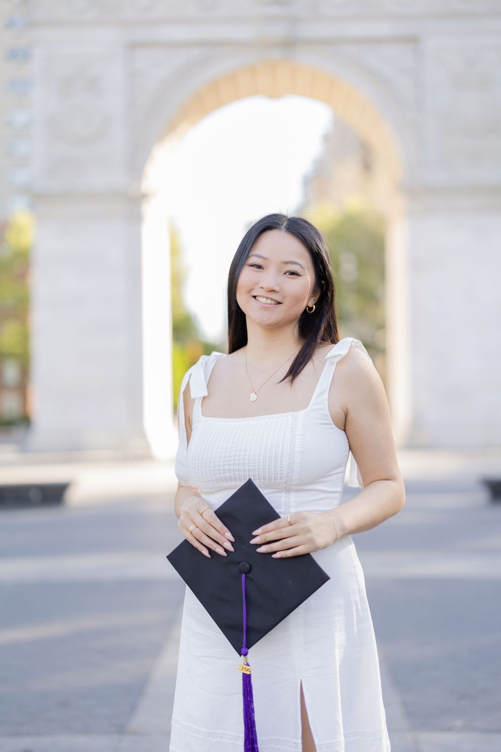 Graduate in white dress holds graduation cap with purple tassel in front of stone archway.