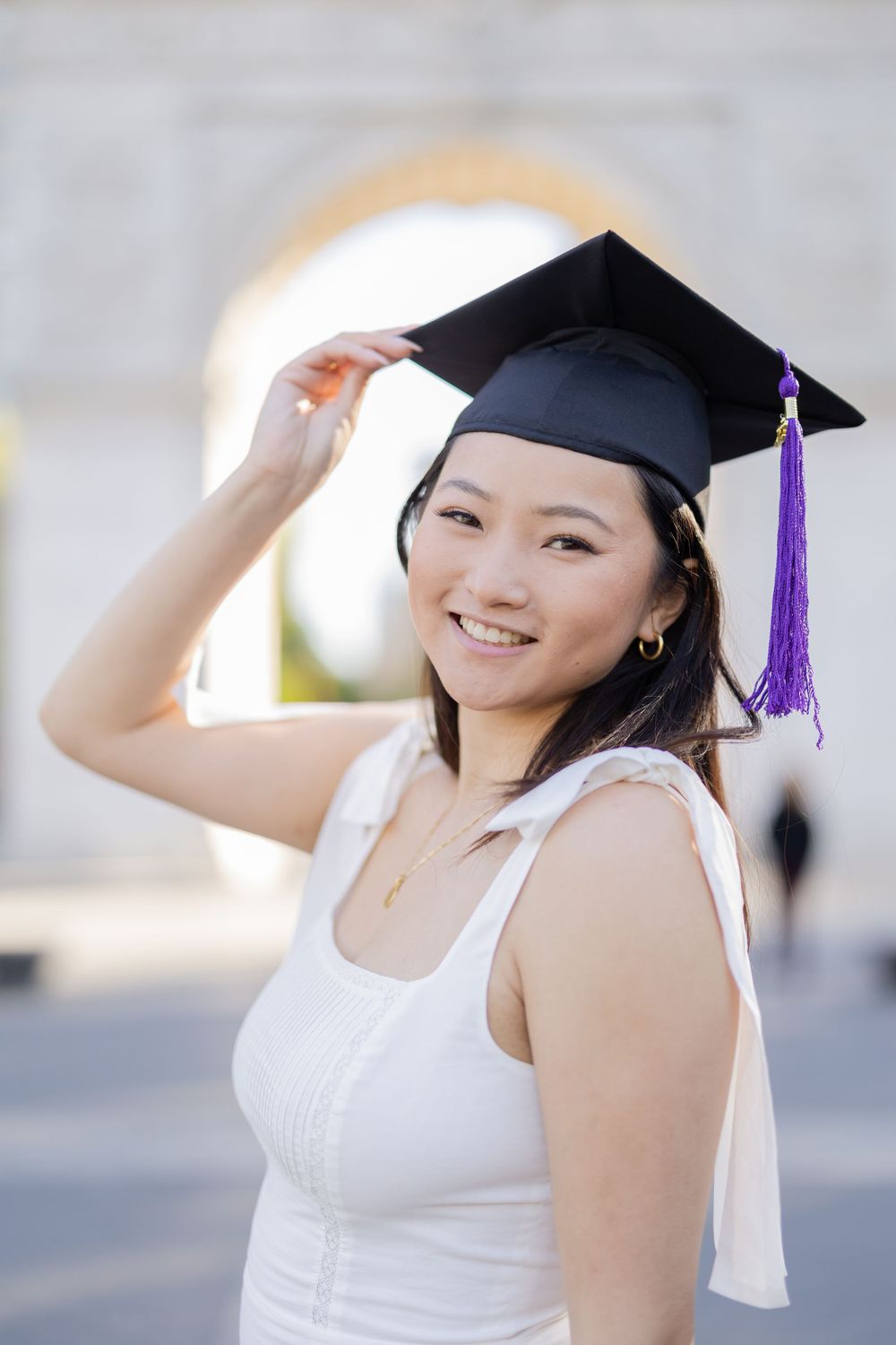 Graduate wearing cap with purple tassel poses in white dress against blurred architecture.