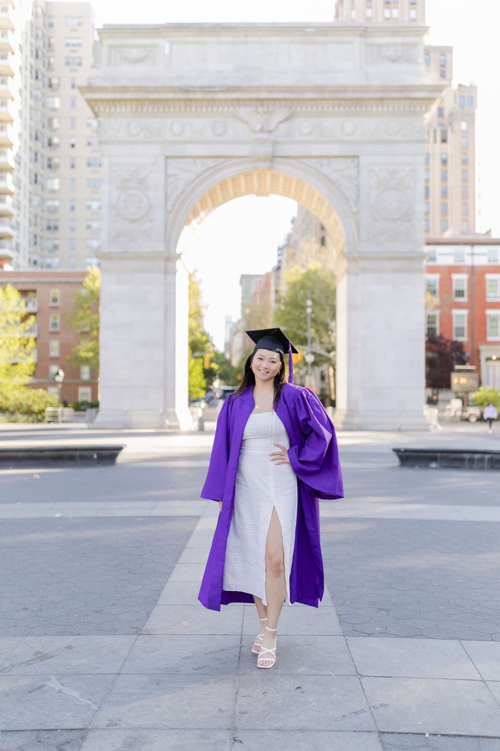 Graduate in purple gown and white dress walks confidently in front of Washington Square Arch.
