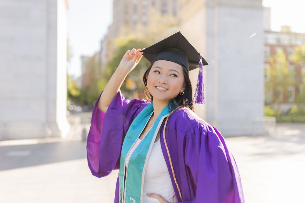 Graduate in purple gown and teal stole poses cheerfully in front of stone architecture.