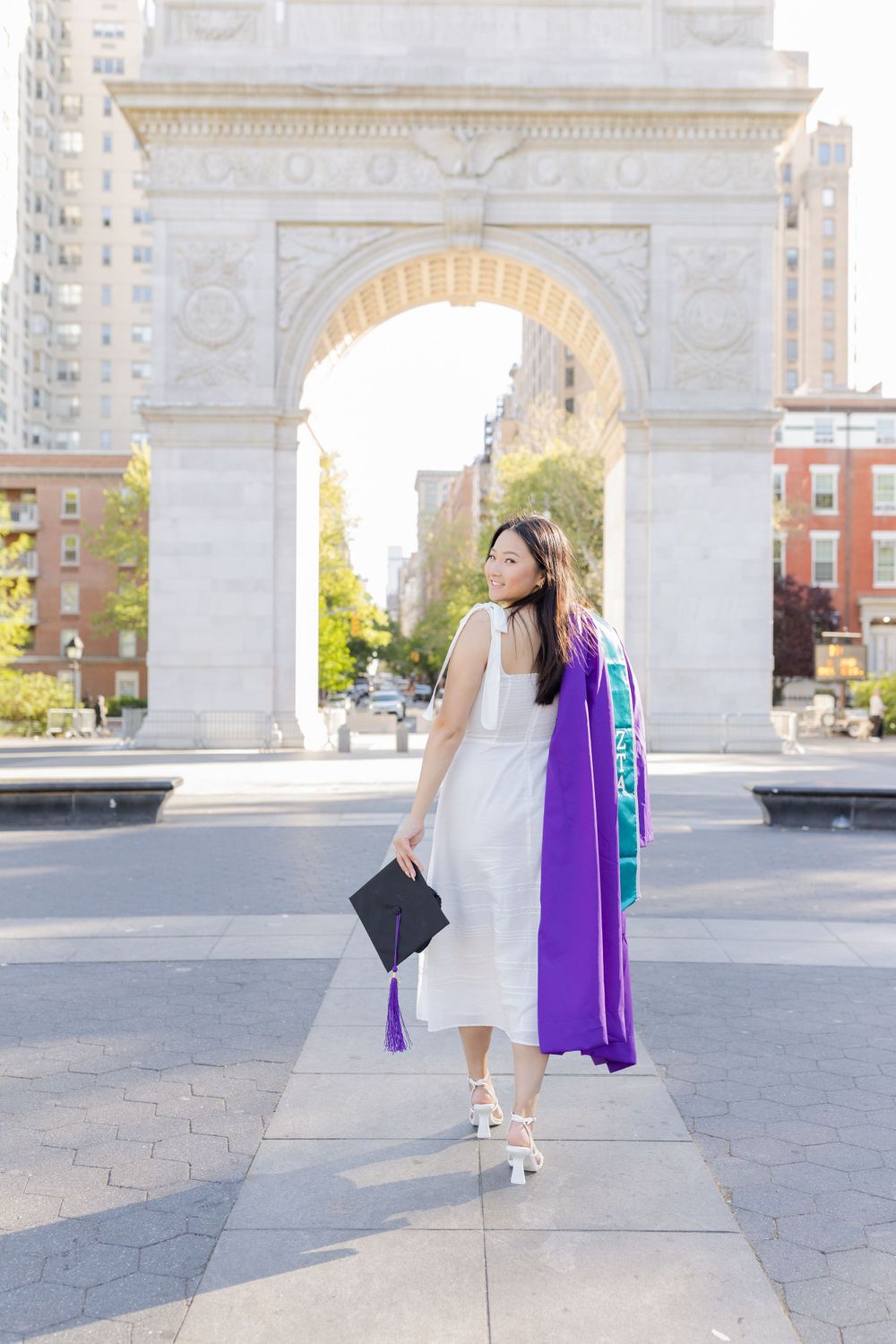 Graduate in white dress and purple gown poses elegantly in front of Washington Square Arch.