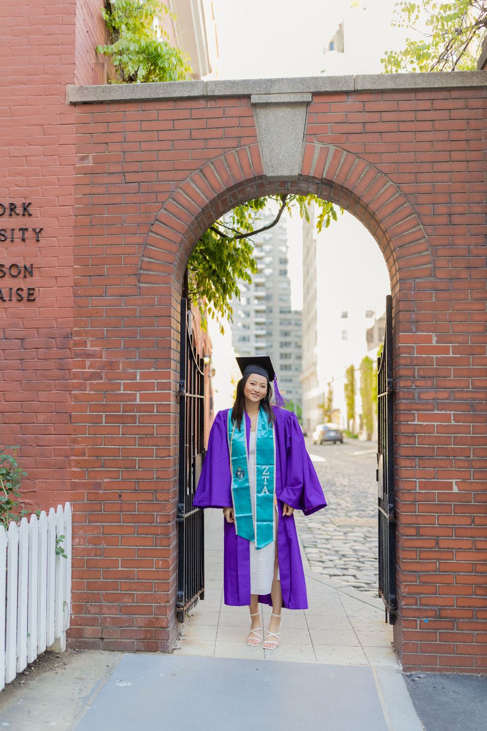 Graduate in purple cap and gown stands under brick archway at New York University.