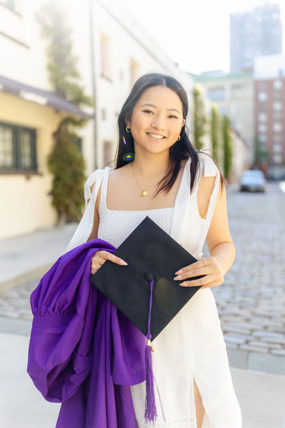 Graduate in white dress holds diploma and purple graduation gown while smiling on cobblestone street.