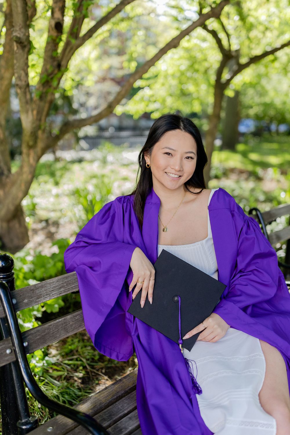 Graduate in purple regalia sits on park bench holding diploma against lush green background.