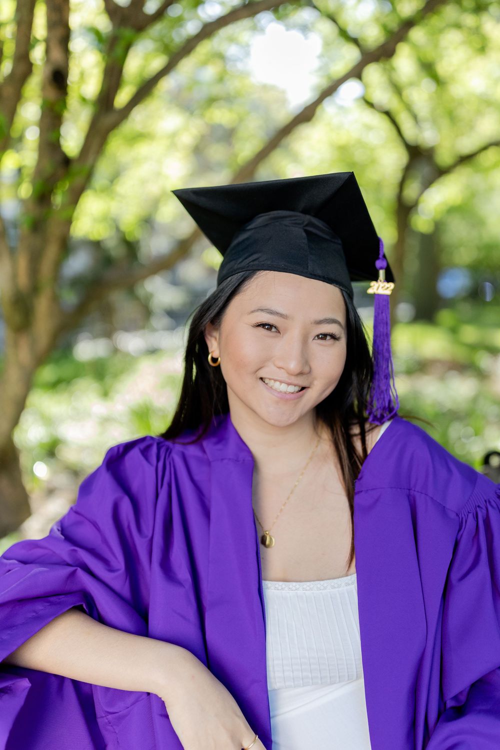 Graduate in purple gown and black mortarboard poses outdoors with trees in background.