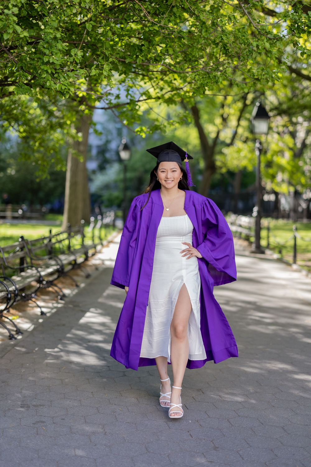 Graduate walks along tree-lined park path wearing purple graduation gown and white dress.