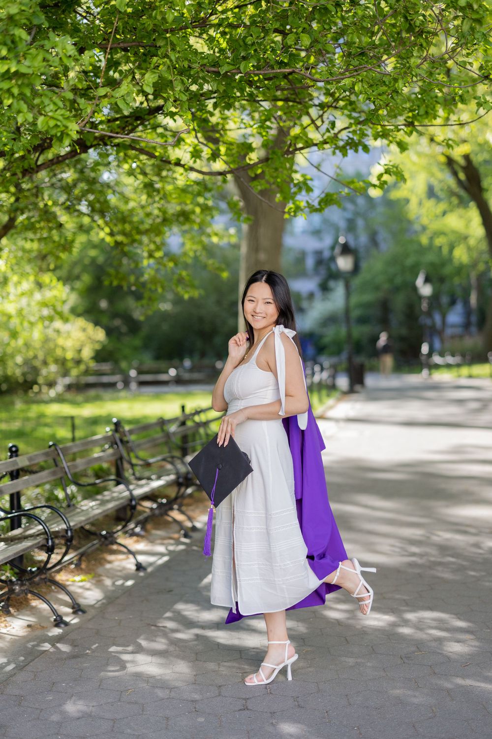 Graduate in white dress carries purple gown and diploma through scenic park pathway.