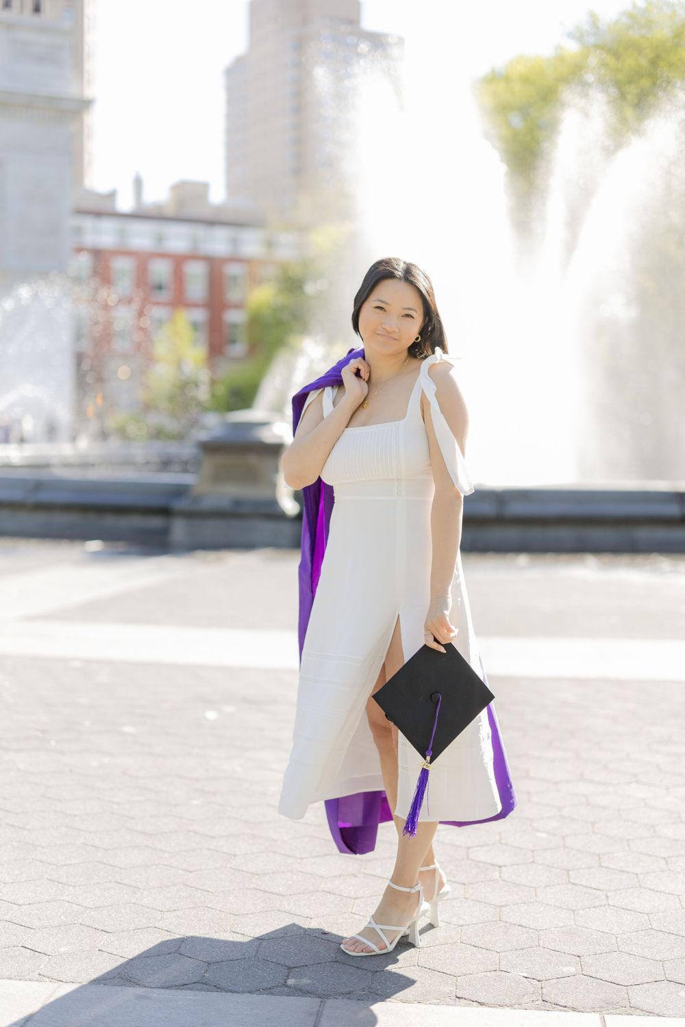 Graduate in white dress holding diploma poses by fountain in bright sunlight.