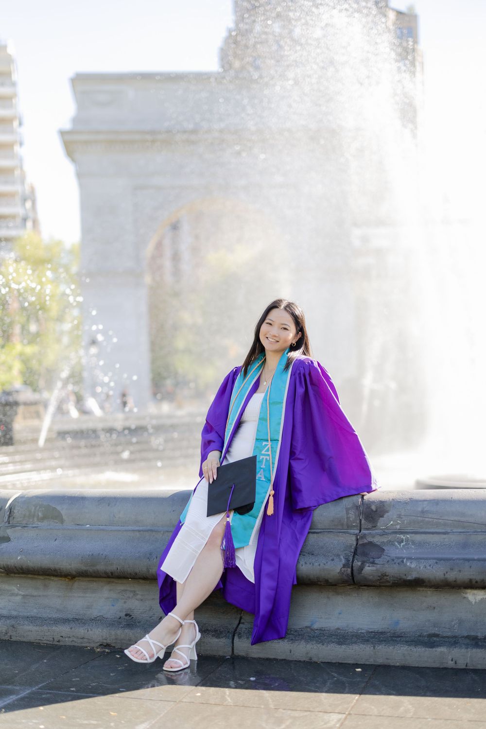 Graduate in purple gown and blue stole poses beside fountain with diploma.