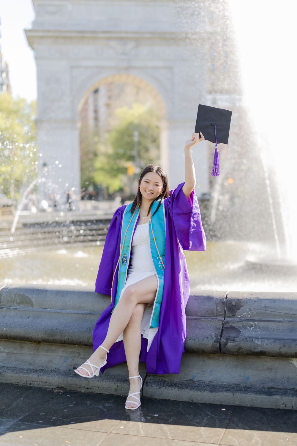 Graduate celebrates by fountain raising diploma while wearing purple graduation regalia.