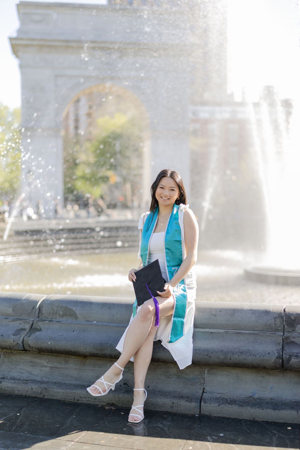 Graduate in turquoise stole and white dress poses by fountain at Washington Square Park graduation photoshoot.