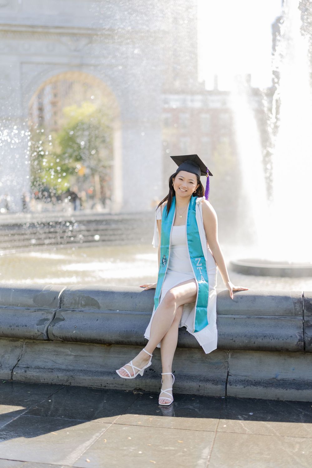 Graduate wearing cap and turquoise stole poses on fountain ledge during sunny graduation photoshoot.