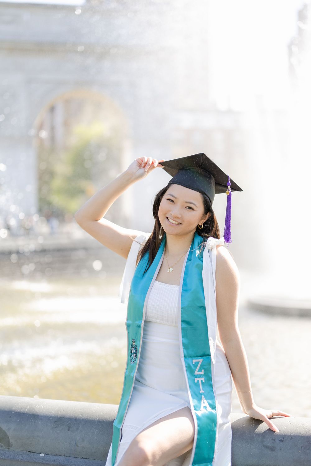 Graduate in turquoise honors stole and mortarboard poses joyfully by fountain during graduation photos.