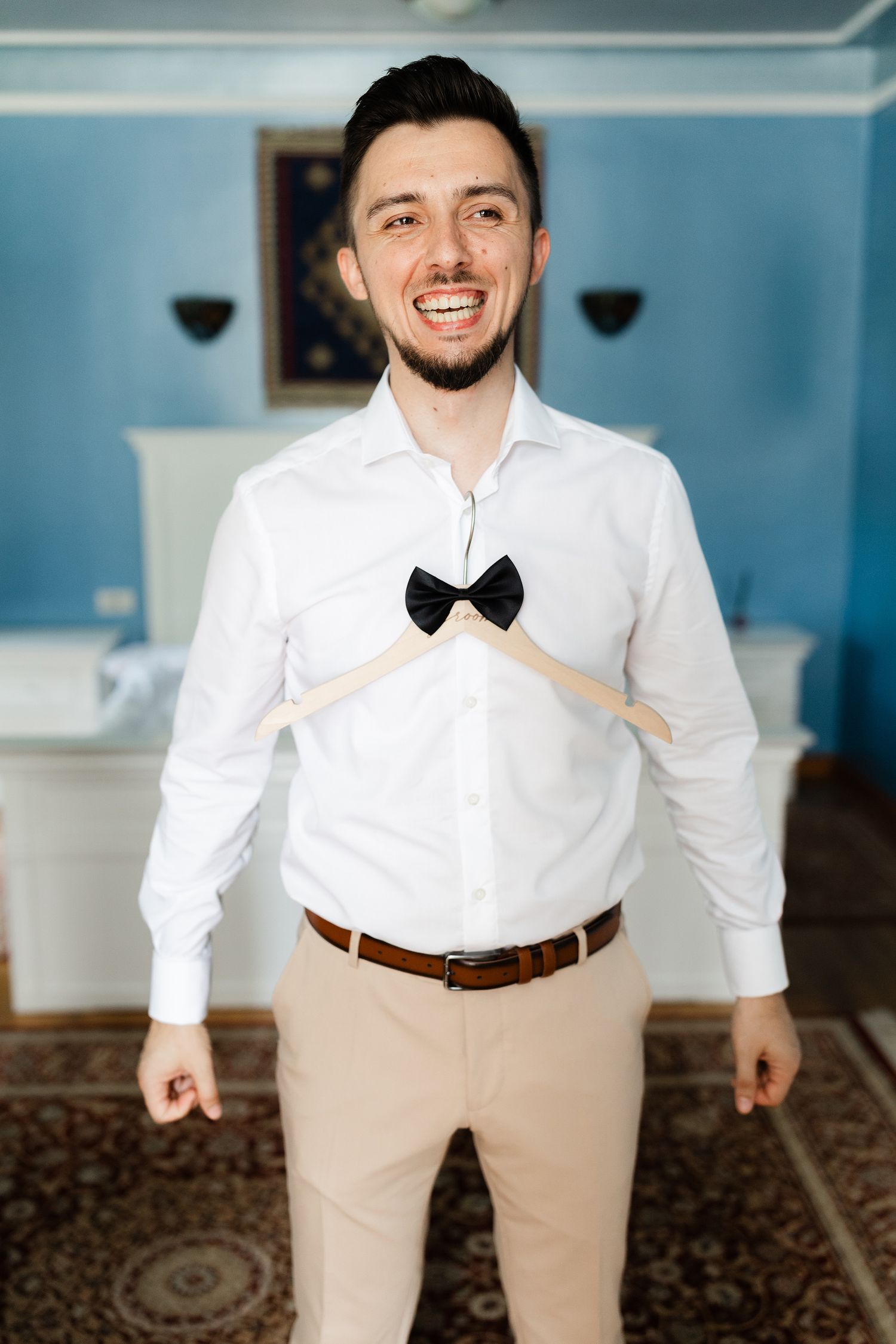 A person in formal attire with a white shirt, khaki pants and bow tie poses in a room with blue walls.