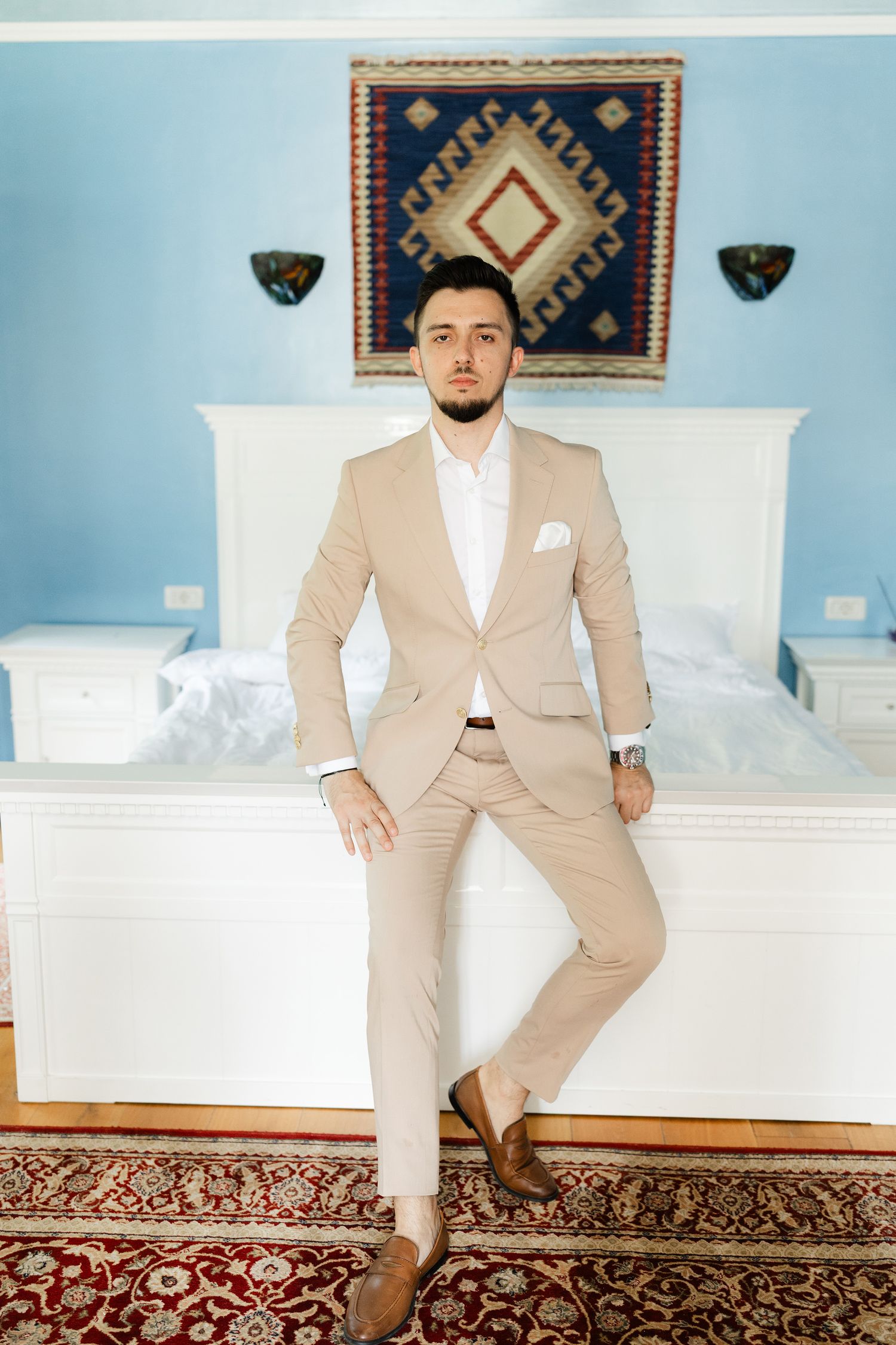 A person in a beige suit and white shirt poses against a blue wall with decorative rugs and artwork.