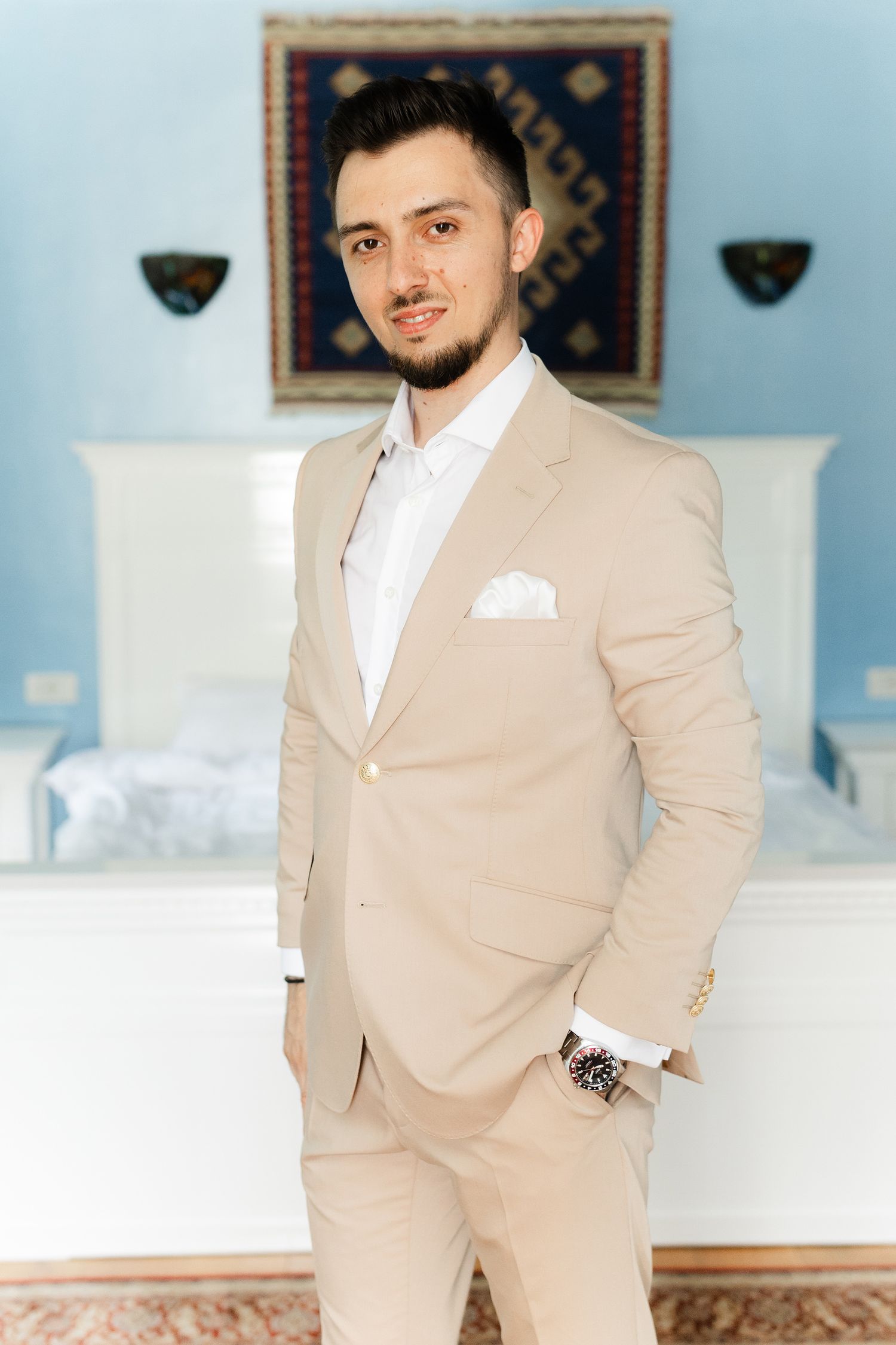A person in a tan suit and white shirt poses in an elegant room with ornate rugs and blue walls.